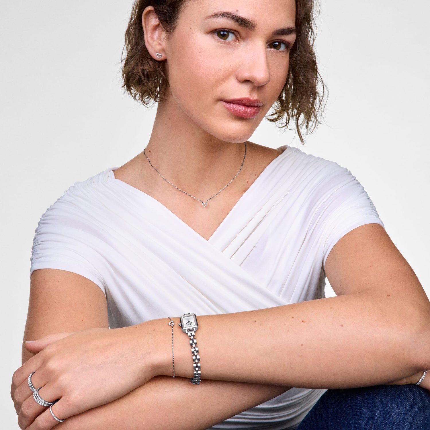 Woman wearing the Mirelle Heart Bracelet Silver with a slim chain and open heart charm set with cubic zirconia stones, styled alongside a silver watch and other jewellery for a polished, everyday look.