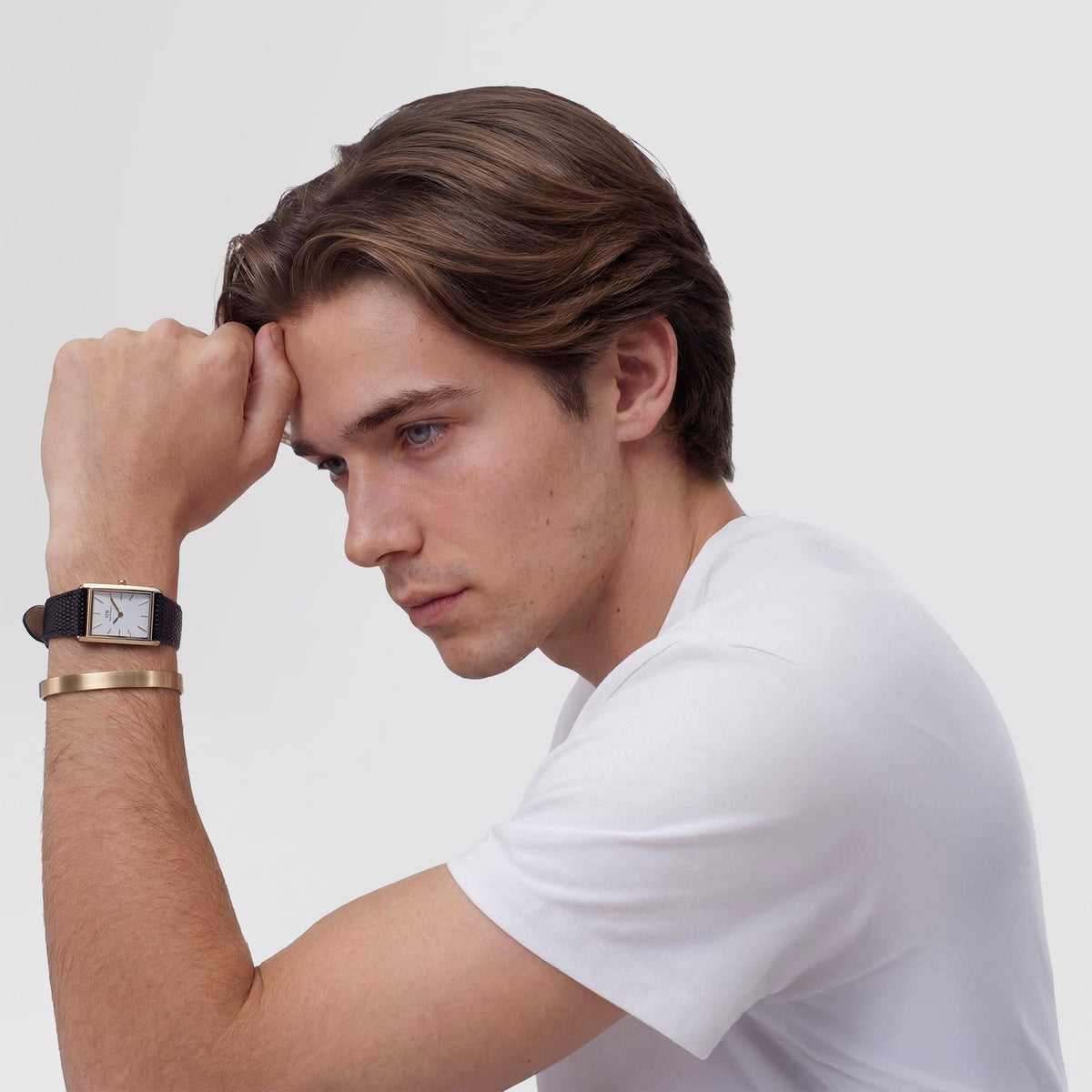 A man wearing a white T-shirt showcases the Bound Black Lizard Guilloché Gold watch on his wrist, highlighting the gold rectangular case, white guilloché dial, and textured black leather strap, styled with a gold-toned bracelet for a modern, everyday look.
