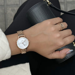 A woman wears the Petite Lumine 5 Link Melrose White MoP Rose Gold watch, featuring a rose gold five-link bracelet and a white mother of pearl dial with crystal markers, styled with a chic ring and a black textured clutch.