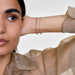 A woman wears the Classic Tennis Bracelet Apricot Silver layered with another bracelet, highlighting the apricot-coloured crystals and slim silver chain against a sheer, neutral blouse.